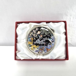May include: A round, silver-toned compact mirror with an Asian-inspired design. The mirror's face features a detailed landscape with deer, cranes, and mountains. The mirror is presented in a red box lined with white satin.