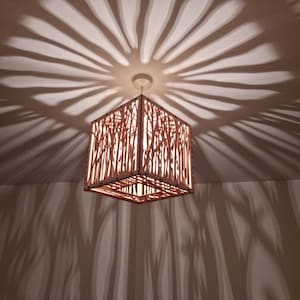 May include: A wooden cube-shaped lampshade with a laser-cut design of branches and leaves. The lampshade casts a shadow of the design on the ceiling.