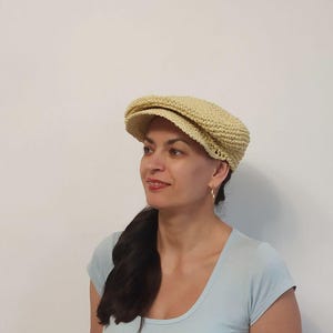 May include: A light beige newsboy cap with a rounded crown and a short, stiff brim. The cap is made of a woven material. The cap is a classic style, suitable for various outfits. The cap is worn by a person.