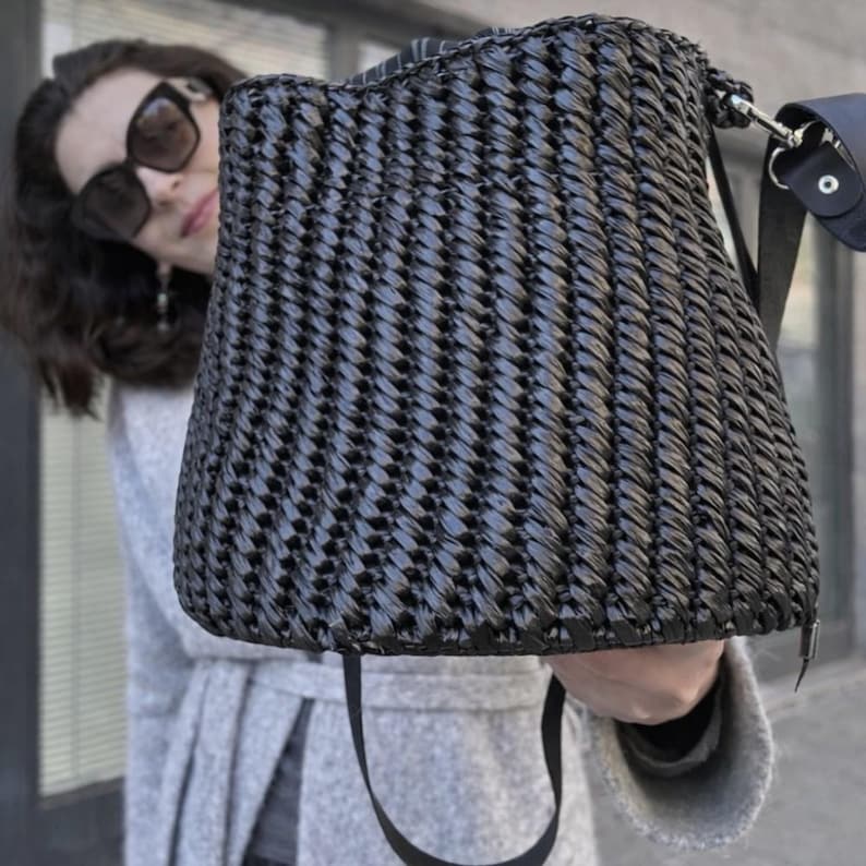 Pu&ograve; includere: Una borsa a secchiello nera con un motivo esterno intrecciato e testurizzato. La borsa presenta una forma strutturata e affusolata, con un cinturino nero fissato lateralmente tramite un anello in metallo argentato e una tracolla abbinata.