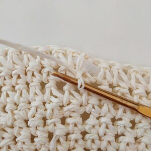 May include: Close-up of a white crocheted fabric with a gold crochet hook. The hook is being used to create a knot in the fabric.