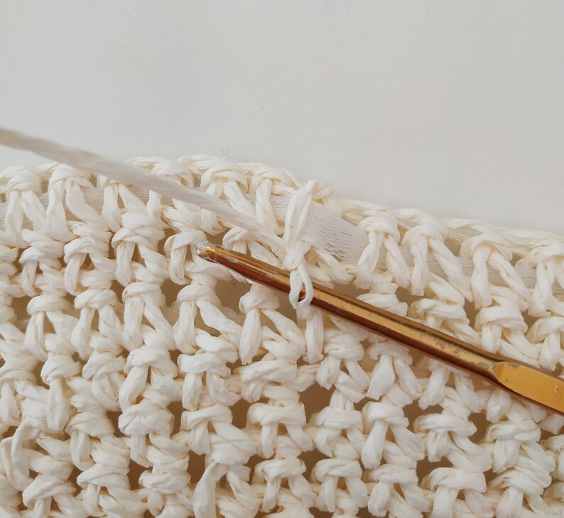 May include: Close-up of a white crocheted fabric with a gold crochet hook. The hook is being used to create a knot in the fabric.