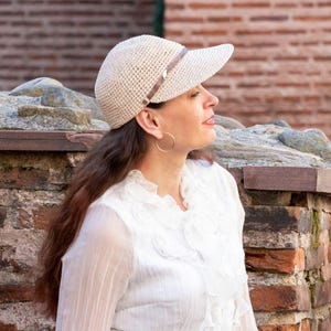 Può includere: Un cappello con visiera all'uncinetto color crema con una fascia marrone. Il cappello ha una tesa curva ed è indossato da una persona con lunghi capelli castani. La persona indossa orecchini a cerchio dorati e una camicetta bianca. Lo sfondo è un muro di mattoni.