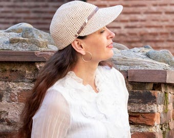 May include: A cream-colored, crocheted visor hat with a brown band. The hat has a curved brim and is worn by a person with long brown hair. The person is wearing gold hoop earrings and a white blouse. The background features a brick wall.