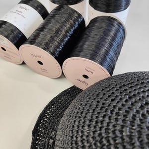 Pu&ograve; includere: Diverse bobine di filato di fibra vegetale di rafia nera Ispie con etichette bianche e un cappello all'uncinetto nero parzialmente visibile. Le etichette delle bobine riportano il testo Ispie, Raffia e 210 Meters, con un motivo a croce.