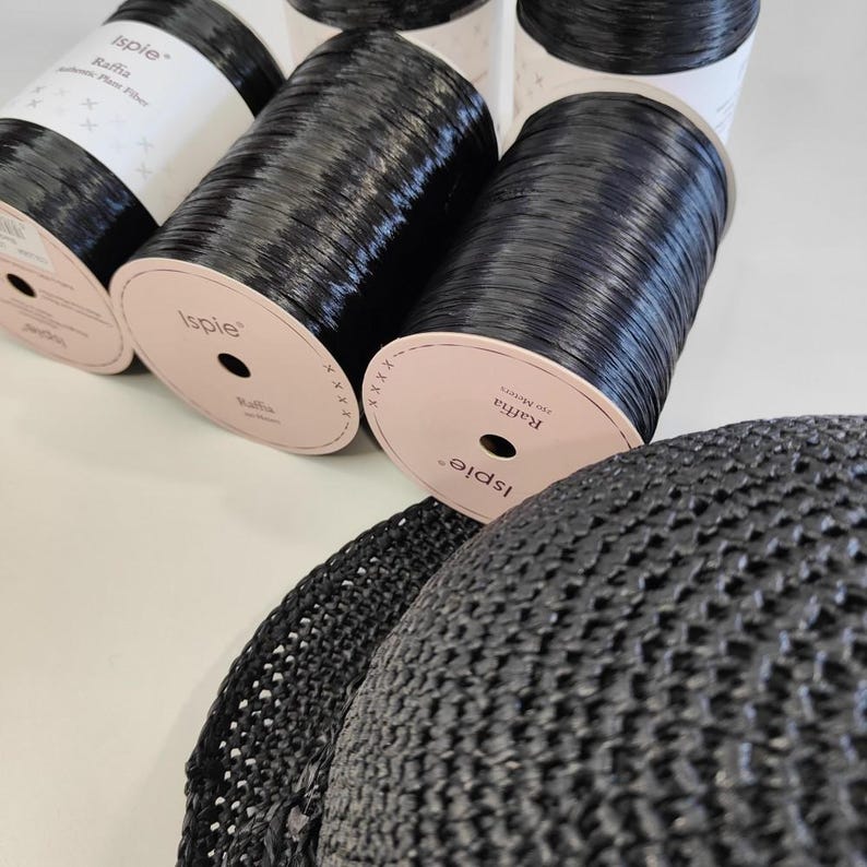 Pu&ograve; includere: Diverse bobine di filato di fibra vegetale di rafia nera Ispie con etichette bianche e un cappello all'uncinetto nero parzialmente visibile. Le etichette delle bobine riportano il testo Ispie, Raffia e 210 Meters, con un motivo a croce.