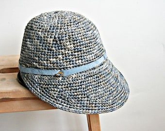 May include: A woven sun hat with a brim and a light blue band. The hat is made of a mix of blue, beige, and white fibers. The hat is resting on a wooden surface.