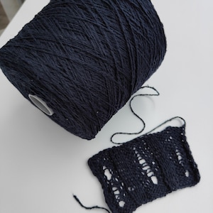May include: A large spool of navy blue yarn next to a small, knitted swatch. The swatch displays an openwork pattern. The yarn is a deep, solid color, and the swatch shows the texture of the knitted fabric.
