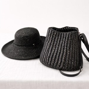 Pu&ograve; includere: Un cappello in paglia intrecciata nera a tesa larga e una borsa a secchiello in paglia intrecciata nera abbinata, caratterizzata da una trama a coste verticali e una tracolla in pelle nera con attacchi in metallo argentato sul lato.