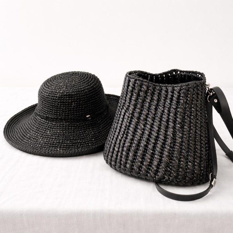 Pu&ograve; includere: Un cappello in paglia intrecciata nera a tesa larga e una borsa a secchiello in paglia intrecciata nera abbinata, caratterizzata da una trama a coste verticali e una tracolla in pelle nera con attacchi in metallo argentato sul lato.