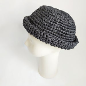 May include: A dark charcoal gray hat with a rounded crown and a short, upturned brim. The hat is constructed from a woven, textured material with a visible interlocking pattern throughout the entire surface, creating a dense and uniform appearance.