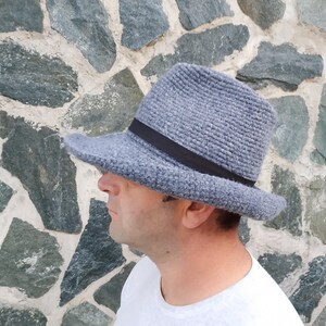 May include: A gray fedora hat with a black band. The hat is made of a textured material and has a classic brim. The hat is worn by a person in a white shirt, set against a stone wall background.