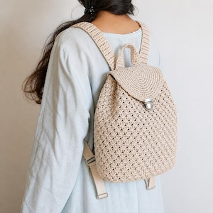 May include: A beige crochet backpack with adjustable straps and a flap closure. The bag features a textured, open weave design and a silver-coloured clasp. The backpack has a top handle and is made of a soft, natural-looking material.