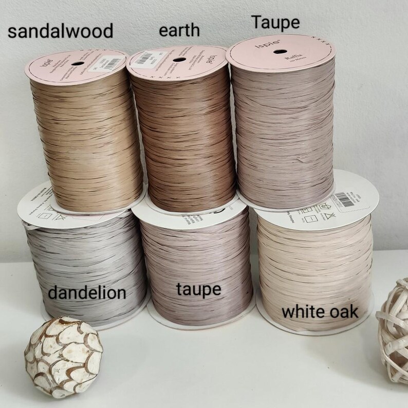 Pu&ograve; includere: Sei rocche di filo di rafia Ispie disposte su due file. La fila superiore presenta i colori etichettati come sandalwood, earth e taupe. La fila inferiore mostra i colori dandelion, taupe e white oak. Ogni rocca ha un'etichetta bianca rotonda.