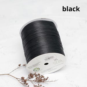 Pu&ograve; includere: Una bobina di nastro in rafia nera realizzato in fibra vegetale. La bobina presenta un'etichetta circolare bianca con testo stampato che include dettagli sul materiale, un codice a barre e istruzioni per la cura. Il nastro ha un aspetto piatto e strutturato.