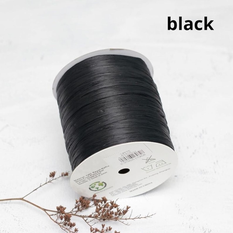 Pu&ograve; includere: Una bobina di nastro in rafia nera realizzato in fibra vegetale. La bobina presenta un'etichetta circolare bianca con testo stampato che include dettagli sul materiale, un codice a barre e istruzioni per la cura. Il nastro ha un aspetto piatto e strutturato.