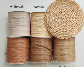 Può includere: Cinque rocche di nastro in rafia nelle tonalità etichettate white oak, oatmeal, autumn, wood e Kraft, disposte con un pezzo circolare in rafia lavorato all'uncinetto. Il nastro ha una finitura opaca e testurizzata con etichette chiare.