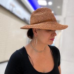 May include: A woven straw fedora hat with a brown leather band. The hat has a wide brim and a classic shape. The hat is worn by a person wearing a black top and silver jewelry.