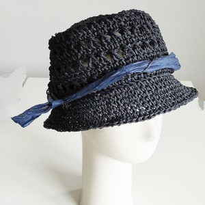 May include: A black crocheted hat with a blue ribbon tied around the crown. The hat is sitting on a white mannequin head.