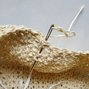 May include: A close-up of a needle and thread being used to sew a beige crocheted hat. The needle is threaded with a white string and is being used to stitch the hat together.