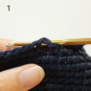 May include: A close-up of a crocheted project in progress. A gold crochet hook with the word "T.BIRDS" is inserted into a stitch. The hook is held by a hand, and the yarn is white.