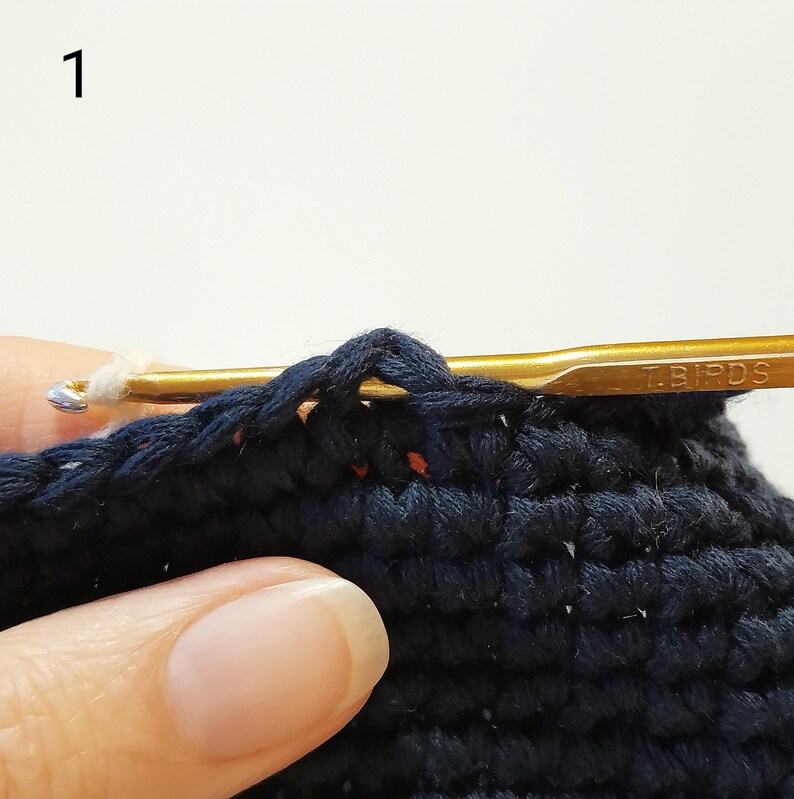 May include: A close-up of a crocheted project in progress. A gold crochet hook with the word "T.BIRDS" is inserted into a stitch. The hook is held by a hand, and the yarn is white.