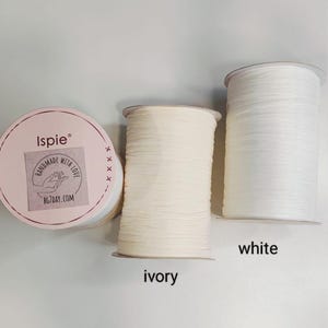Pu&ograve; includere: Tre rocche di filo di rafia Ispie. Una rocca presenta un'etichetta rotonda rosa con il testo Ispie, Handmade with Love e BG7DAY.COM. Le altre due rocche sono cilindriche, una di colore avorio e una di colore bianco brillante.