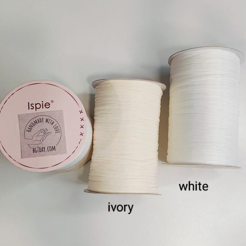 Pu&ograve; includere: Tre rocche di filo di rafia Ispie. Una rocca presenta un'etichetta rotonda rosa con il testo Ispie, Handmade with Love e BG7DAY.COM. Le altre due rocche sono cilindriche, una di colore avorio e una di colore bianco brillante.
