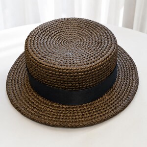 Pu&ograve; includere: Un cappello di paglia marrone scuro modello canotier con corona piatta e tesa circolare larga. Il cappello presenta un nastro in gros-grain nero liscio e solido avvolto attorno alla base della corona, in contrasto con la paglia intrecciata.