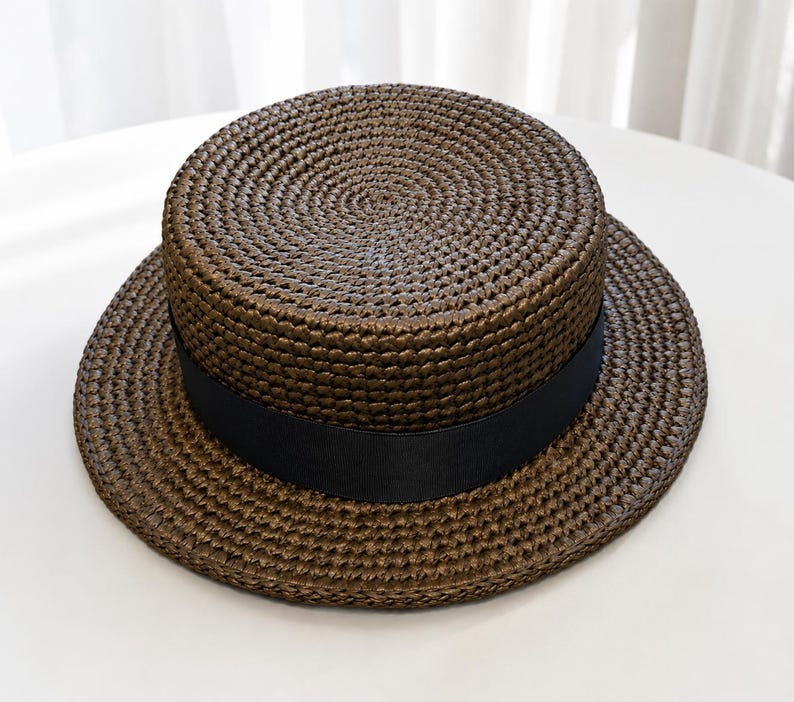 Pu&ograve; includere: Un cappello di paglia marrone scuro modello canotier con corona piatta e tesa circolare larga. Il cappello presenta un nastro in gros-grain nero liscio e solido avvolto attorno alla base della corona, in contrasto con la paglia intrecciata.