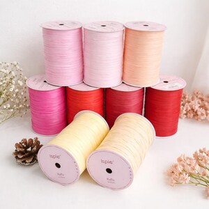 May include: A collection of nine Ispie brand raffia yarn spools arranged in three rows. The spools feature various colors including light pink, soft peach, vibrant magenta, bright red, and pale yellow. Each spool has a circular label.