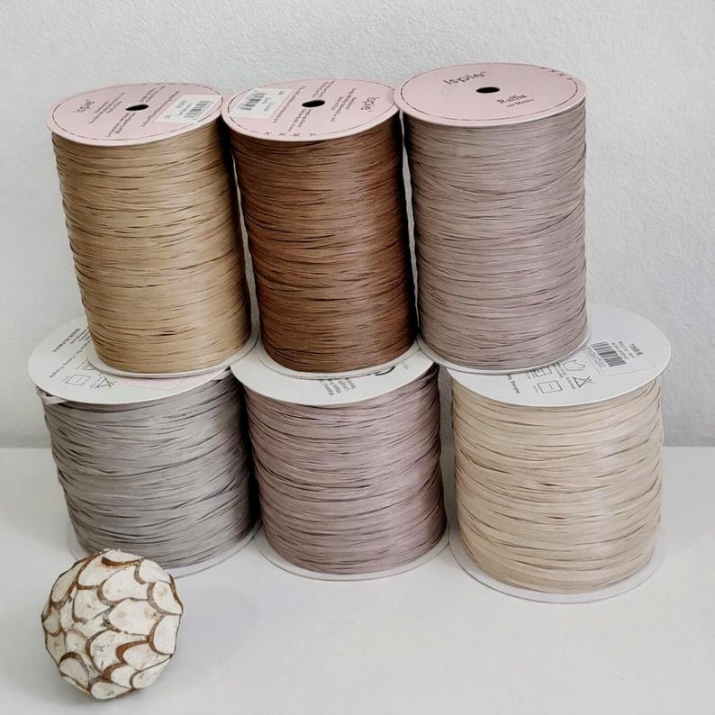 Pu&ograve; includere: Sei bobine cilindriche di nastro in rafia in varie tonalit&agrave; neutre della terra, tra cui marrone chiaro, marrone, tortora e beige, disposte su due file. Un piccolo oggetto decorativo sferico con un motivo a scaglie bianco e marrone &egrave; in primo piano.