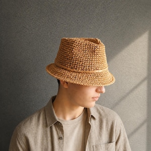 Könnte beinhalten: Ein Fedora-Hut aus geflochtenem Stroh mit einem braunen Lederband. Der Hut hat eine strukturierte Krone und eine leicht abgewinkelte Krempe. Der Hut ist hellbraun und scheint handgefertigt zu sein. Der Hut ist ein klassischer Stil, geeignet für Sonnenschutz.