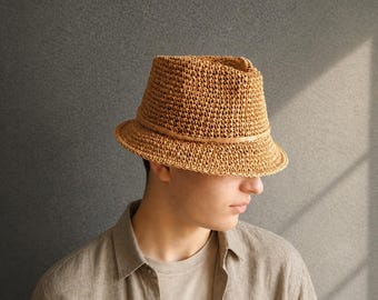 May include: A woven straw fedora hat with a brown leather band. The hat has a structured crown and a slightly angled brim. The hat is a light brown color and appears to be handmade. The hat is a classic style, suitable for sun protection.