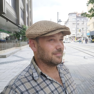 Puede incluir: Una gorra de newsboy de crochet de color beige con una visera plana. La gorra está hecha de un material tejido y tiene una superficie ligeramente texturizada.