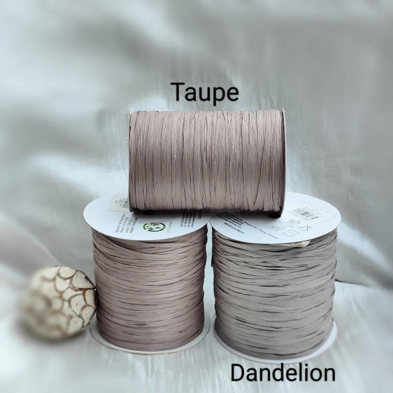 Pu&ograve; includere: Tre bobine di nastro piatto disposte in una pila. La bobina superiore &egrave; etichettata Taupe e la bobina in basso a destra &egrave; etichettata Dandelion. Il nastro ha una consistenza opaca simile alla carta e una larghezza costante, avvolto strettamente su nuclei di plastica bianca.