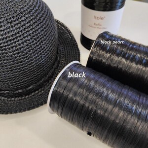 Pu&ograve; includere: Un cappello all'uncinetto nero accanto a due bobine di fibra di rafia nera. Una bobina &egrave; etichettata black e l'altra black pearl. Le bobine sono cilindriche con una consistenza liscia e lucida, e il cappello presenta un fitto motivo intrecciato.