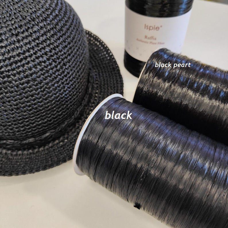 Pu&ograve; includere: Un cappello all'uncinetto nero accanto a due bobine di fibra di rafia nera. Una bobina &egrave; etichettata black e l'altra black pearl. Le bobine sono cilindriche con una consistenza liscia e lucida, e il cappello presenta un fitto motivo intrecciato.