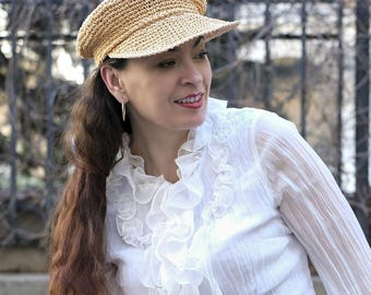 May include: A light brown, crocheted newsboy hat with a short brim. The hat is made of a woven material and has a rounded crown. The hat is worn by a person wearing a white blouse with ruffled details.