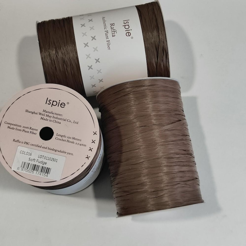 Pu&ograve; includere: Due rocche di filato di rafia Ispie marrone in fibra vegetale autentica. Le etichette indicano 100% rayon, 250 metri di lunghezza e una misura di uncinetto di 2,5-4 mm. Il colore &egrave; Soft Fudge, lotto 01102501, con codice a barre e certificazione FSC.