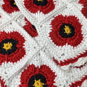 May include: A white crochet blanket featuring red, black and yellow flowers. The blanket is made using a granny square pattern.