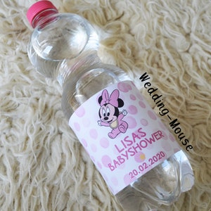 以下が含まれることがあります： ピンクのキャップが付いた透明なプラスチックウォーターボトル。ラベルには、ピンクのドットと「LISAS BABYSHOWER 20.02.2020」の文字が入ったミニーマウスの漫画デザインが描かれています。ボトルは白くふわふわした表面にあります。
