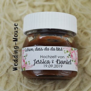 Könnte beinhalten: Ein kleines Glas gefüllt mit Schokoladenaufstrich, mit einem weißen Deckel und einem Etikett mit der Aufschrift "Schön, dass du da bist" und "Hochzeit von Jessica & Daniel 19.09.2019". Das Etikett hat ein florales Design mit rosa Rosen.