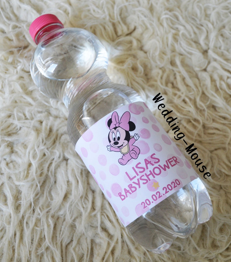 以下が含まれることがあります： ピンクのキャップが付いた透明なプラスチックウォーターボトル。ラベルには、ピンクのドットと「LISAS BABYSHOWER 20.02.2020」の文字が入ったミニーマウスの漫画デザインが描かれています。ボトルは白くふわふわした表面にあります。