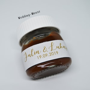 Könnte beinhalten: Ein kleines Glas gefüllt mit brauner Streichmasse, mit einem weißen Etikett, auf dem "Julia & Lukas 19.09.2019" in goldenen Buchstaben steht.