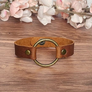 以下が含まれることがあります： 大きな真鍮色の円形の金属リングが付いた茶色の革のブレスレット。ブレスレットには、真鍮色の小さな金属製の留め金が2つ付いています。