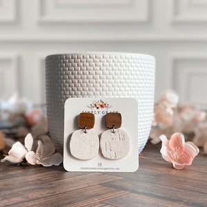 May include: A pair of white and brown square earrings with a wood-like texture. The earrings are displayed on a white card with the text "Simply Grace" and a website address.
