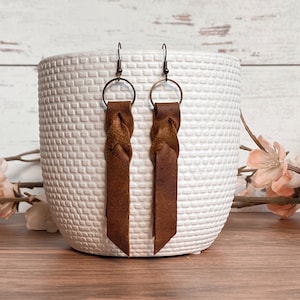 以下が含まれることがあります： 茶色の革製イヤリング。上部に編み込みのディテールがあり、細長い長方形の形をしています。イヤリングはブロンズ色のフープとフックから吊り下げられています。イヤリングは、背景にピンクの花がある白いテクスチャード加工のベースの前に展示されています。
