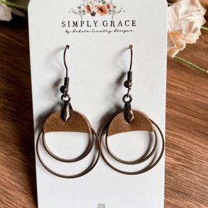 May include: A pair of brown leather earrings with double gold hoops. The earrings are on a white card with the text "SIMPLY GRACE by Dakota Country Designs" and "dakotacountrydesigns.etsy.com".