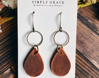 Peut inclure: Une paire de boucles d'oreilles en forme de goutte d'eau en cuir marron avec un anneau doré et un crochet doré. Les boucles d'oreilles sont sur une carte blanche avec le texte "SIMPLY GRACE by dakota country designs" et "dakotacountrydesigns.etsy.com".